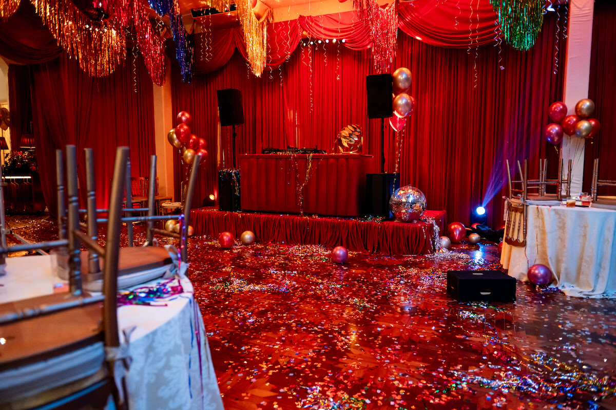 Um salão em tons de vermelho onde uma festa acabou de terminar tem papel picado e adereços brilhantes por toda parte