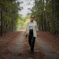 Woman in sunglasses and casual clothes walking down a forest path holding a handgun, surrounded by tall pine trees, with a red structure visible in the background.