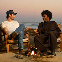 Two people sitting in wooden chairs by a campfire on a sandy beach at sunset, engaged in conversation with the ocean and pastel sky in the background.