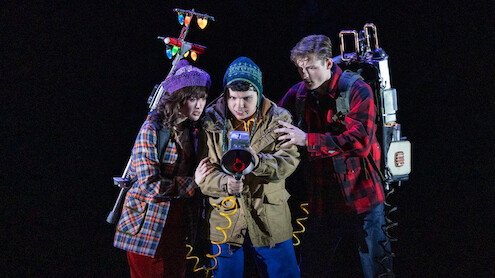 Three young people in winter clothes, holding ghost-busting equipment with glowing lights, stand closely together in a dark, dramatic setting, appearing focused and ready for action.