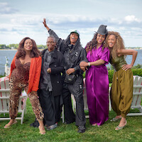 Five women posing and smiling outdoors by a lake, with Adirondack chairs on grass, all wearing colorful, expressive outfits under a partly cloudy sky.