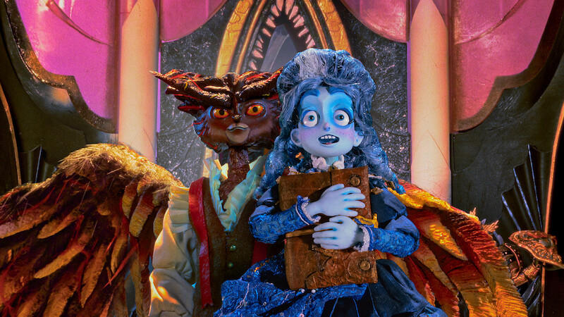A blue-skinned, animated girl in a blue dress holds a book, standing nervously in front of a menacing owl-like creature with wings inside a lavish, ornately decorated room with pink and gold arches and dramatic lighting.