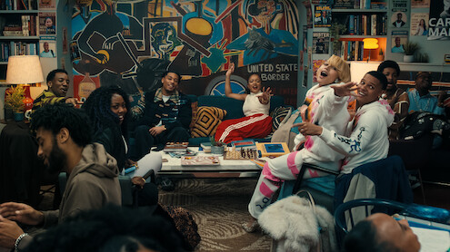 Um grupo diverso de pessoas ri e conversa em uma sala de estar colorida repleta de obras de arte, livros e um grande mural, criando uma atmosfera vivaz e receptiva.