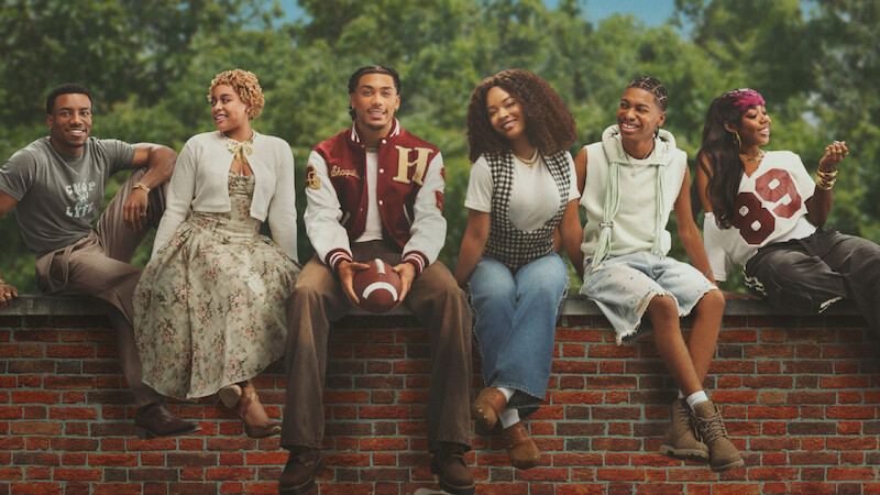 Six young adults sit on a brick wall outdoors, smiling and relaxed. Trees and blue sky in background. One holds a football; they wear casual, varied outfits, indicating a friendly, laid-back group setting.