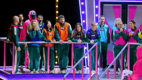 A group of contestants wearing numbered tracksuits stand on a brightly lit game show set, appearing tense and focused. Neon lights and a futuristic, colorful environment add excitement and intensity to the scene.