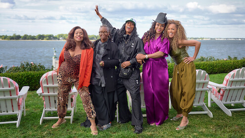 Five stylish women stand outdoors on green grass in front of white chairs by a lake, smiling and posing happily under a partly cloudy sky.