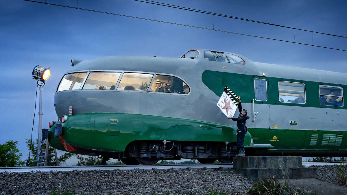 A vintage green and gray streamlined train is stopped outdoors at dusk on train tracks; a person in uniform stands beside it holding a clapperboard while a film light illuminates the scene, creating a cinematic atmosphere.