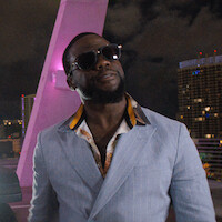 Man in sunglasses and a light blue suit stands confidently on a rooftop at night, with city lights and modern buildings in the background, illuminated by purple lighting.