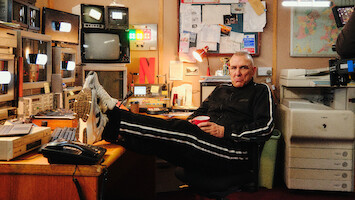 Vinnie Jones in a tracksuit relaxes with feet on desk in a cluttered control room, surrounded by monitors, electronics, bulletin board, and office equipment, holding a red cup.