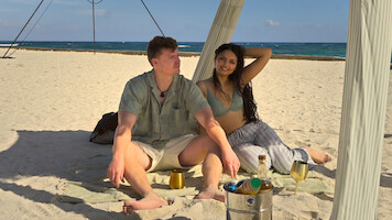A man and woman relaxing on a beach under a canopy, sitting on a blanket with drinks and a bucket of beverages, with the ocean and blue sky in the background.