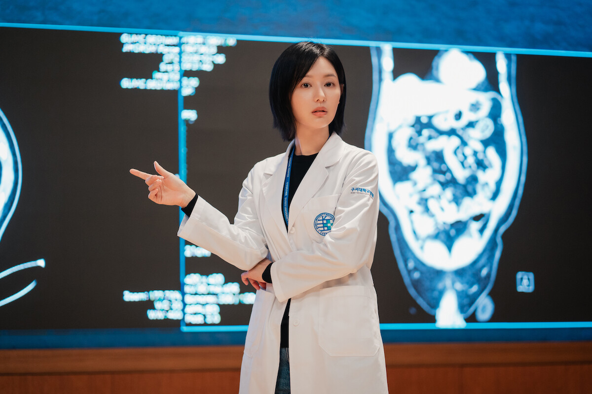 Uma mulher sul-coreana veste jaleco branco e aponta para uma projeção médica na tela