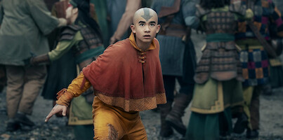 A young person in orange and yellow monk-style clothing with a blue arrow tattoo on their head stands alert in a crowded outdoor setting with people in armor, suggesting a tense or urgent situation.
