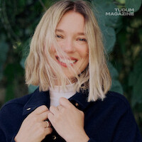 Smiling woman with shoulder-length blonde hair stands outdoors wearing a navy coat and white turtleneck, hands near collar, with green foliage background and "TUDUM MAGAZINE" logo in upper right corner.