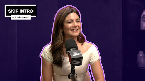 Monica Barbaro smiles in front of a microphone, framed by a deep purple background with a matching purple outline surrounding her.