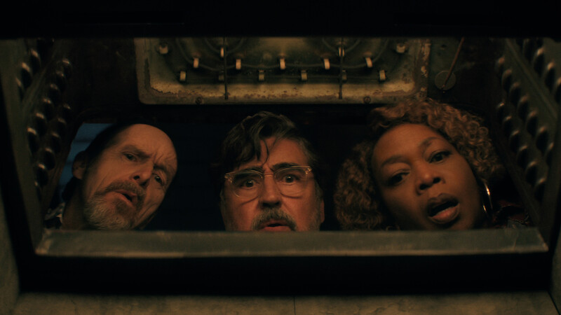 Three adults, two men and one woman, peer with curiosity and surprise into an open oven, as seen from inside the oven looking out in a dimly lit kitchen setting in 'The Boroughs.'