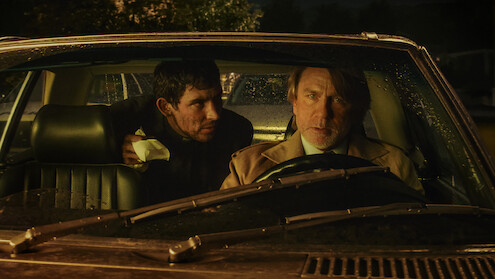 Two men sit in a car at night, illuminated by dim streetlights and rain on the windshield. The driver looks concerned while the passenger leans in with urgency, creating a tense and suspenseful atmosphere.