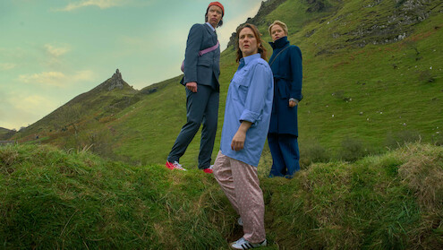 Three people stand on grassy hills under a cloudy sky, dressed in modern, colorful clothes, with a mountain peak in the background, creating a contrast between natural landscape and urban fashion.