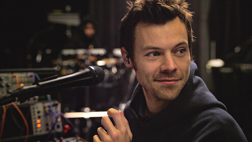 Harry Styles in a hoodie sits at a microphone with electronic equipment around him in a dimly lit studio or rehearsal space, looking off to the side and smiling slightly.