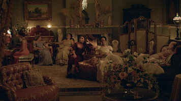 Women in elegant period dresses sit and lounge in a richly decorated parlor with vintage furniture, ornate decor, flowers, and a lit fireplace, creating a warm, historical atmosphere.