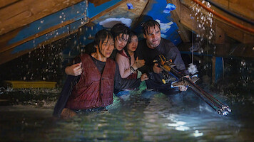 Four people waist-deep in floodwater in an attic, huddled together, appearing worried and alert, with one person holding a weapon, water pouring from ceiling, scene is tense and dramatic.