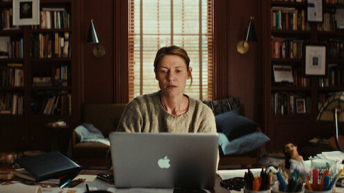 A woman sits at a cluttered desk in a cozy, book-filled home office, focused on her laptop. Warm light and wooden bookshelves create a thoughtful, slightly tense, and introspective mood.
