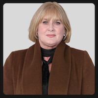 A woman with short blonde hair wearing a brown coat and black scarf poses against a plain light gray background in a studio or professional setting.
