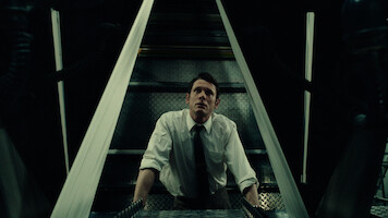 Man in white shirt and tie looks upward in a dimly lit industrial or mechanical setting, framed by metallic structures, suggesting a tense or suspenseful atmosphere.