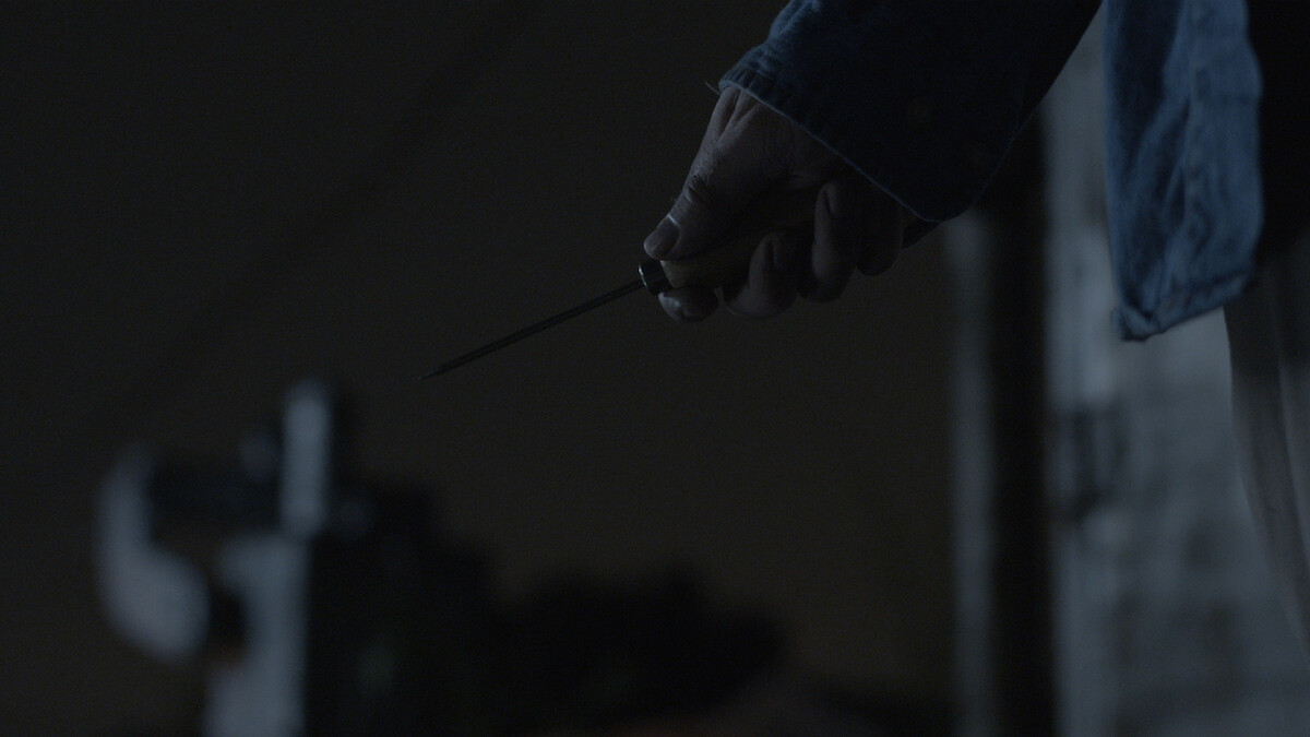 A person holding a knife in their right hand at night, wearing a dark blue jacket. The background is dimly lit, creating a suspenseful or tense atmosphere.