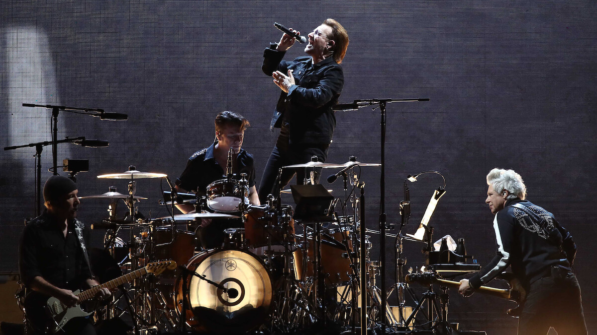 A rock band performs on stage with dramatic lighting; the lead singer stands on the drum riser singing into a microphone as other band members play guitar, drums, and keyboard in a concert setting.