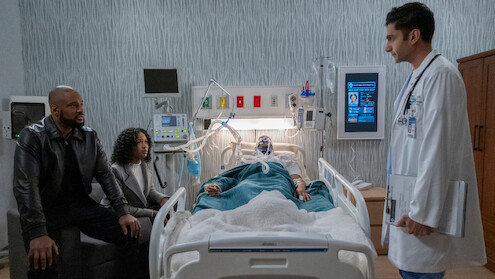 A patient lies in a hospital bed connected to medical equipment, with a man and woman seated nearby and a doctor standing at the bedside in a modern hospital room.