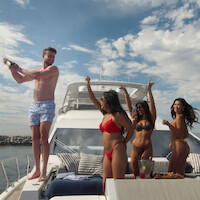 A man in swim trunks opens champagne while three women in bikinis celebrate on the deck of a luxury yacht, under a partly cloudy sky, with drinks and snacks nearby, dock and water visible in the background.